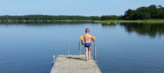 Ett barn med simpuffar på armarna står på en badbrygga vid en sjö.